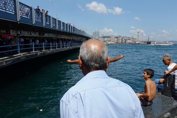 Elderly man watching boys near water by a bridge, capturing a street coincidence in an urban waterfront setting.