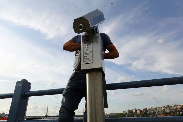 A man aligns perfectly behind a street coin-operated binocular, creating a witty street coincidence in an urban setting.