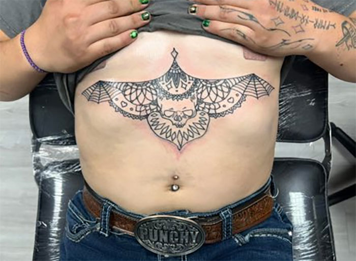 Black ink tattoo of a bat with webbed wings and skull design placed across upper stomach and ribs.