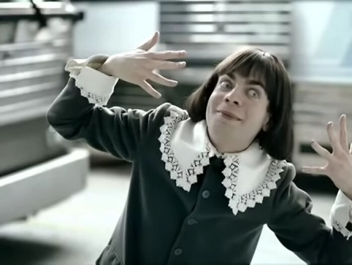 Person in a creepy commercial making an eerie facial expression and hand gestures in a dimly lit setting.