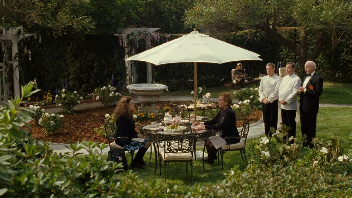 Two women having tea in a lush garden setting with attendants nearby, evoking mysterious magical imaginary worlds.