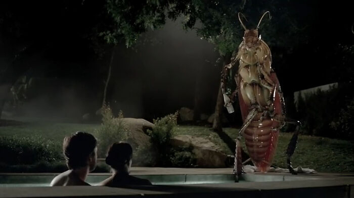 Two people in a pool at night staring at a giant creepy insect figure in a dark, eerie outdoor setting.