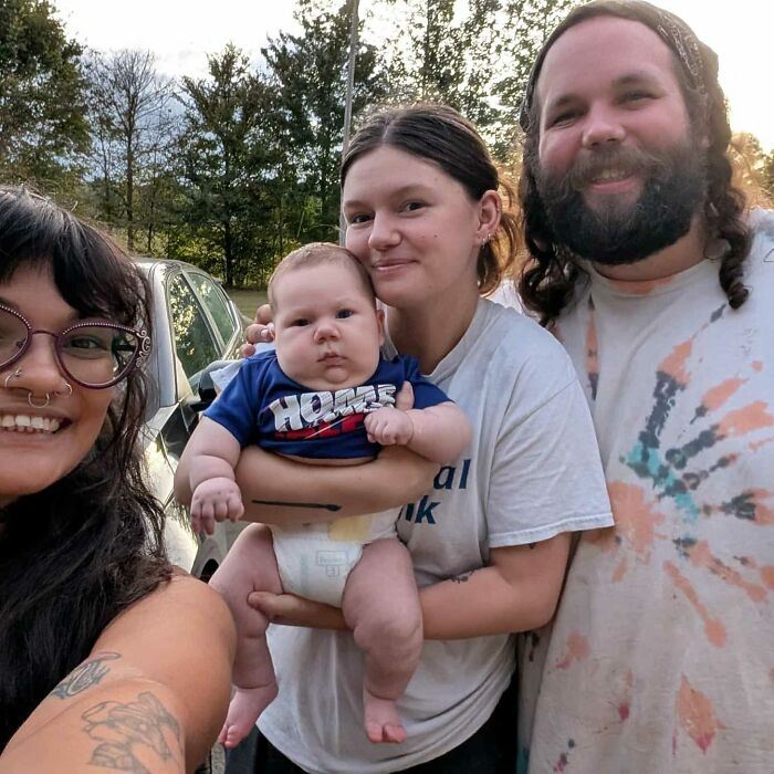 A mom holding her baby who broke hospital record, smiling with two adults outdoors near a car in daylight. A mom holding her baby who broke hospital record, smiling with two adults outdoors near a car in daylight.