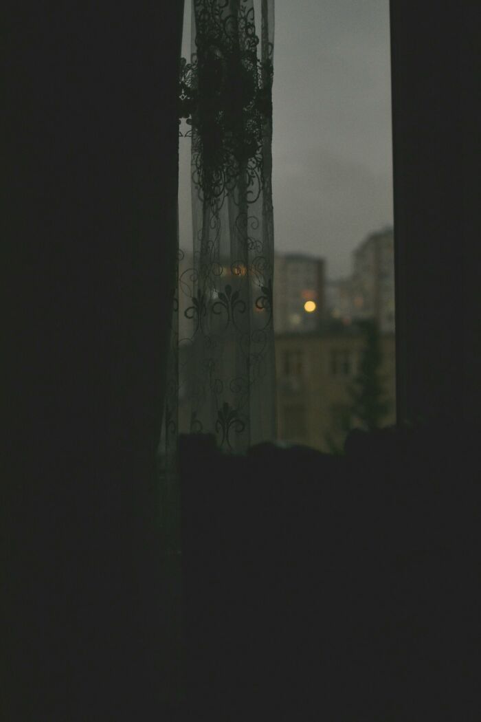 Dark window scene with lace curtain and faint city lights outside, evoking a creepy Halloween-inspired mood for a would you rather poll.