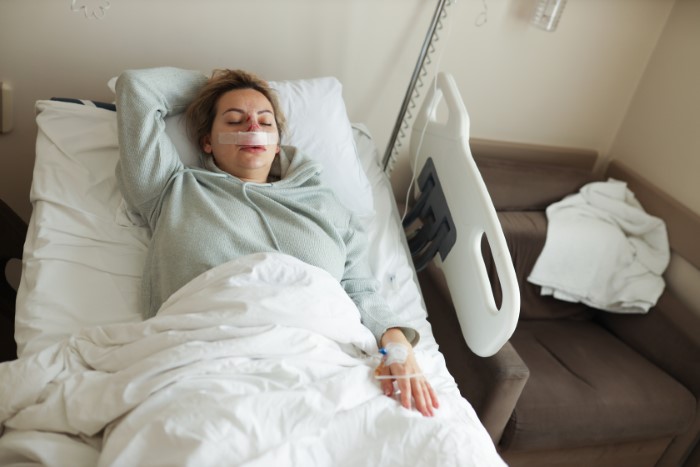 Woman resting in hospital bed with nasal bandage and IV, relating to doctor refusing to rehire sister after costly mistake.