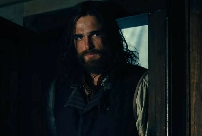 A man with long hair and beard in period costume standing in a dimly lit room from Wuthering Heights trailer.