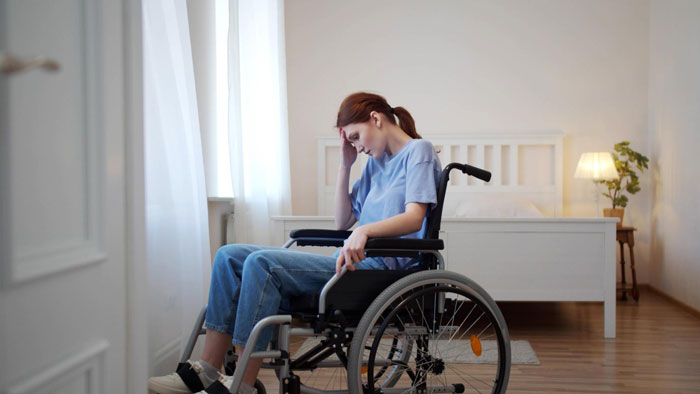 Teen disabled girl sitting in a wheelchair looking upset and shocked in a bedroom with natural light from window.