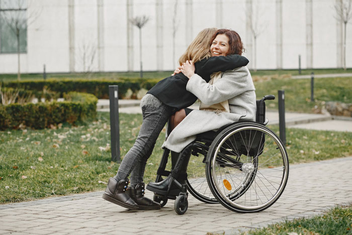Disabled teen in wheelchair sharing a warm hug with her best friend outdoors on a paved path near a garden.