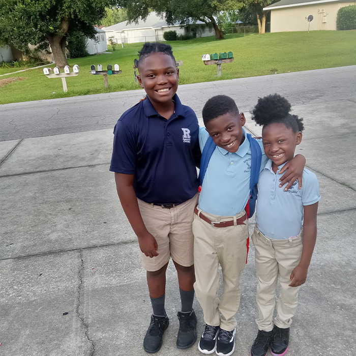 Three smiling children in school uniforms standing outside on a driveway, related to Netflix&rsquo;s The Perfect Neighbor true story.