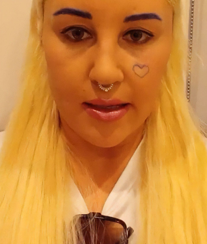 Amanda Bynes with bright blonde hair, blue eyebrows, septum piercing, and heart tattoo on cheek in close-up selfie.