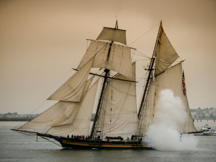 Tall ship sailing on the ocean with sails fully open and a large plume of smoke near the bow on a calm day.