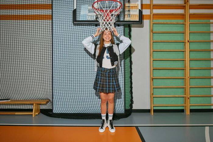 Teen girl in school gym wearing plaid skirt and jacket, standing under basketball hoop, representing popular kids in school.