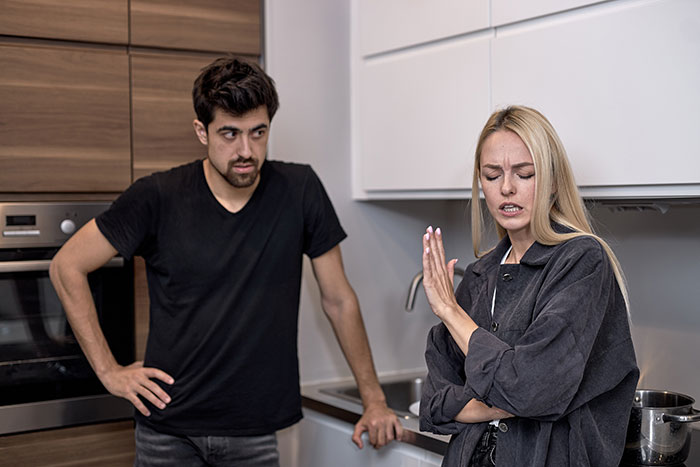 Wife standing upset with arms crossed as husband looks on in kitchen, highlighting struggles with bed cleaning and refusal to use aid.