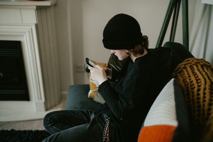 Teen sitting on couch wearing black hat and clothes, focused on phone screen, representing popular kids in school behavior.