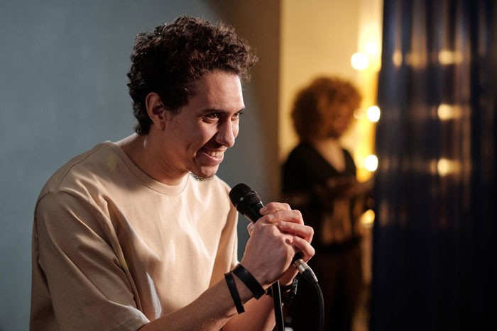 Young man smiling while holding a microphone indoors with a blurred woman in the background, capturing the annoying toxic friend photographer vibe.