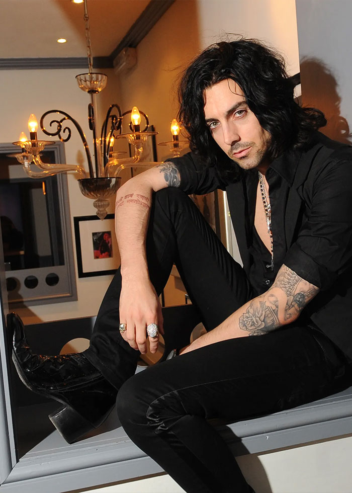 Rock star with tattoos and black outfit posing indoors, linked to fatal prison attack and crimes against children case.