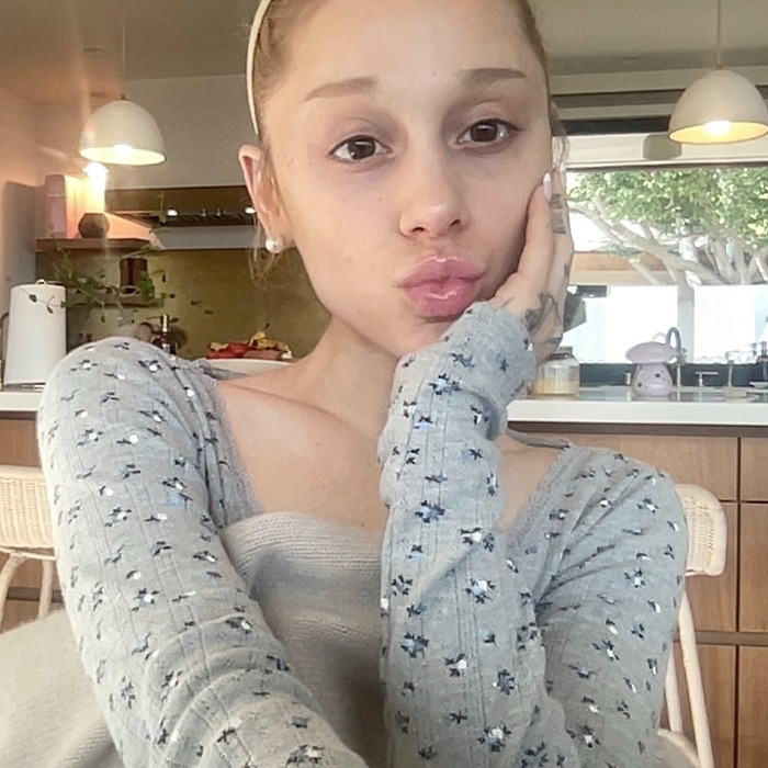 Ariana Grande with a dramatic new look, posing indoors, wearing a patterned grey top, facing fan backlash online.