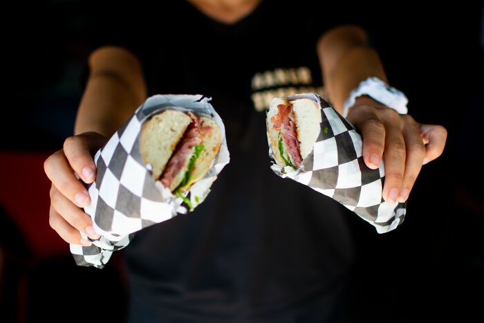 Close-up of a person holding a sandwich bite split in half wrapped in black and white checkered paper with a dark background.