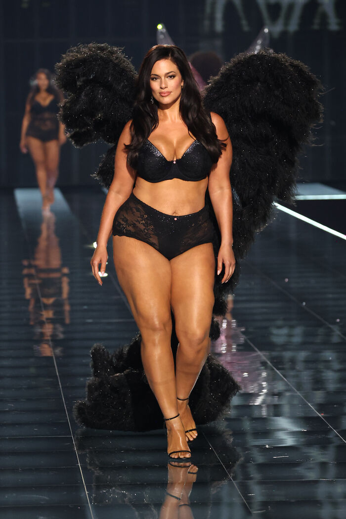 Plus size model confidently walking runway in black lingerie with large feathered wings at Victoria's Secret fashion show 2025.