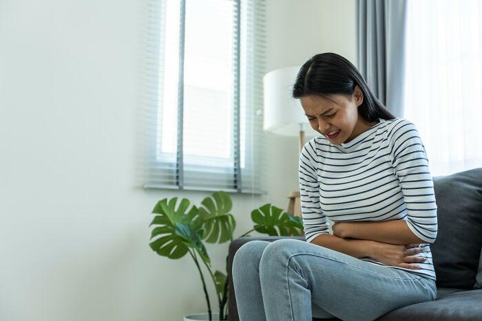 Woman sitting indoors clutching stomach in pain, illustrating medical problems that are far worse than people think.