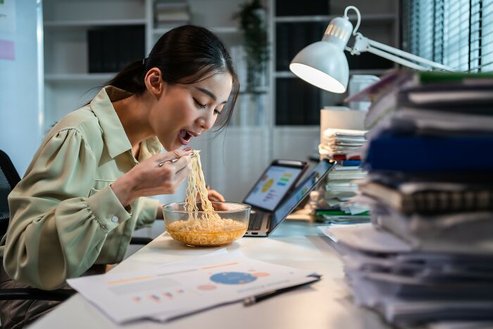 Young woman eating noodles at a desk cluttered with papers and laptop, highlighting busy lifestyle over healthy habits.