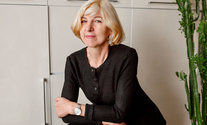 Mature woman with blonde hair wearing a black top, posing indoors with arms crossed near a tall green cactus plant.
