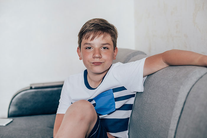 11-year-old boy sitting on a gray couch with a neutral expression acting beyond creepy around stepsister who gave birth