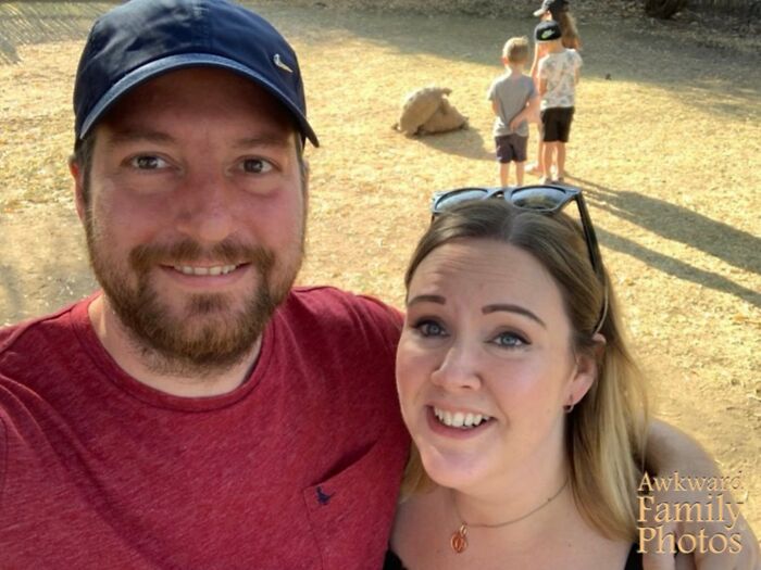 Pareja sonriente en selfie con niños y oveja de fondo, foto familiar incómoda icónica al aire libre en días soleados.