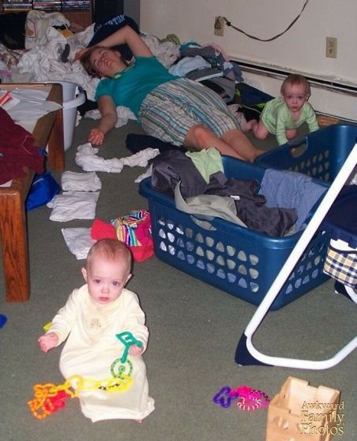 Tired mom lying on the floor amid laundry baskets and babies with toys in a chaotic family photo moment.