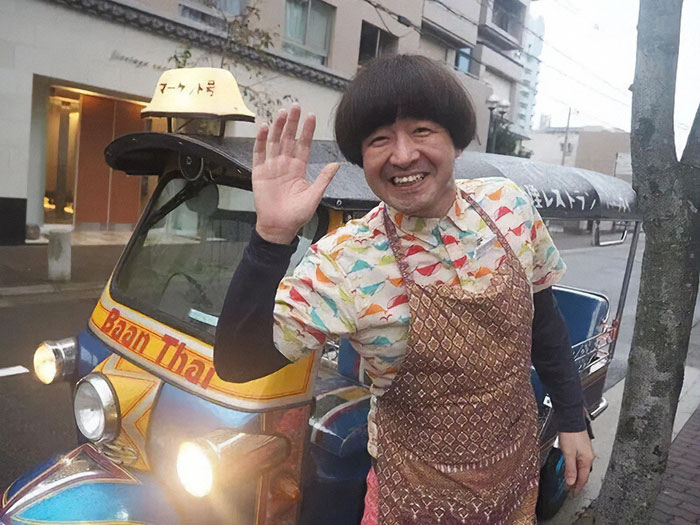 Man in apron waving near colorful tuk-tuk on street, related to tourists kicked out of Japanese restaurant debate. Man in apron waving near colorful tuk-tuk on street, related to tourists kicked out of Japanese restaurant debate.