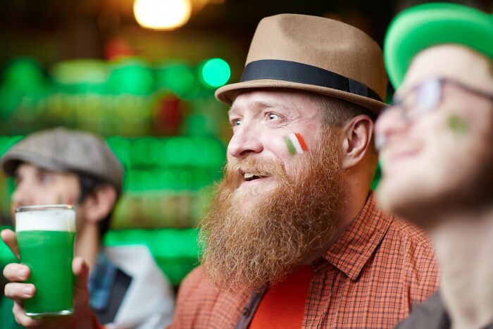 Bearded man with Irish flag face paint holding green beer, celebrating positive stereotypes about various countries in a lively pub.