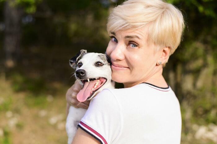 Woman smiling happily while holding her intelligent pet dog outdoors in a sunny natural setting.