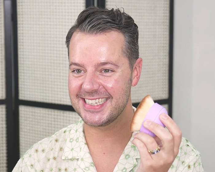 Man smiling and holding a makeup brush demonstrating application in relation to Victoria's Secret models fake tan controversy.