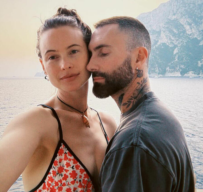 Victoria&rsquo;s Secret model posing with a tattooed man by the sea with mountains in the background on a sunny day.