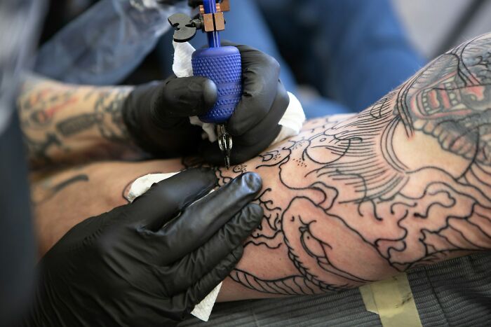 Tattoo artist wearing black gloves using a blue tattoo machine to ink a detailed design on a client's arm.