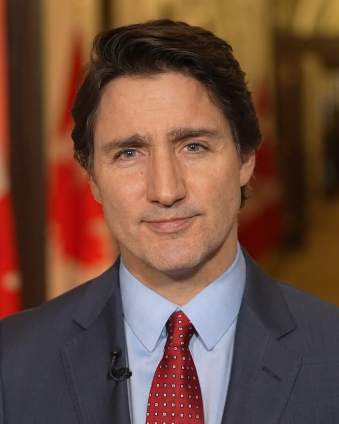 Justin Trudeau in a formal suit with red tie, posing indoors with blurred Canadian flags in the background.