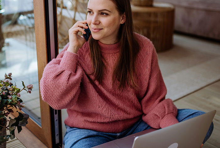 Young woman in a cozy pink sweater discovering loopholes while talking on the phone and using a laptop at home.