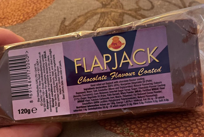 Hand holding a chocolate flavour coated flapjack snack bar with label showing ingredients and nutrition details.