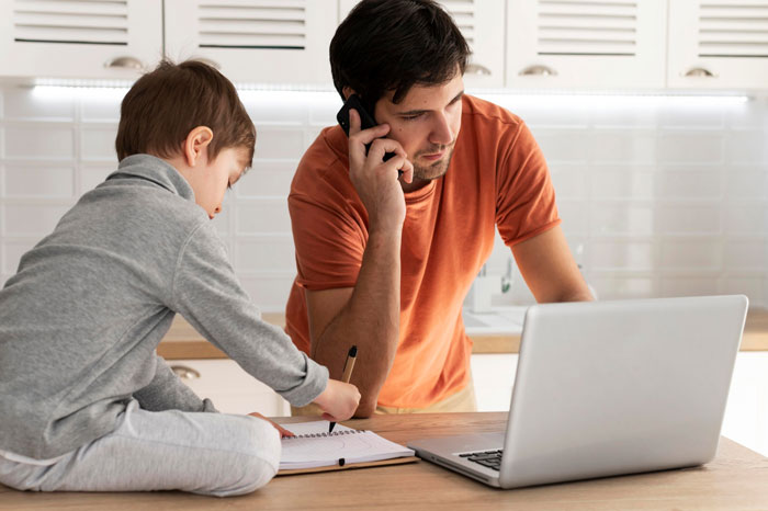 Man on phone with laptop nearby while young boy writes in notebook on kitchen table, planning wedding travel and budget concerns. Man on phone with laptop nearby while young boy writes in notebook on kitchen table, planning wedding travel and budget concerns.