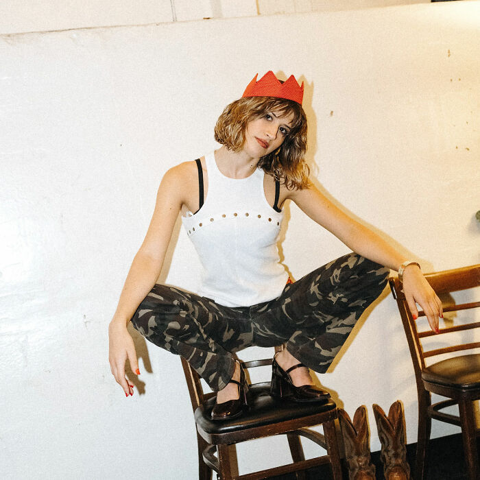 Young woman wearing a red paper crown and camo pants posing on a chair embodying the Group 7 TikTok viral trend.