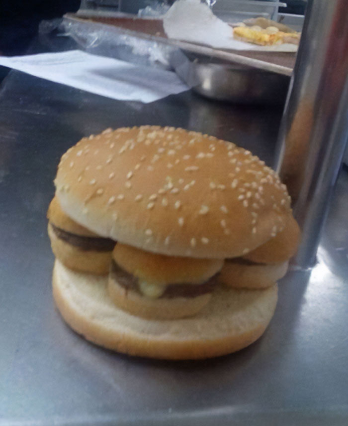 Weird and cursed burger with multiple mini sliders inside a sesame seed bun creating a strange food mashup on a metal surface.