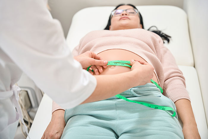 Patient lying on a bed while a medical professional measures the abdomen during a botched plastic surgery consultation.