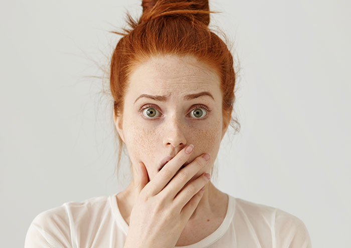 Red-haired woman with green eyes covering her mouth, showing shock and concern about botched plastic surgeries.