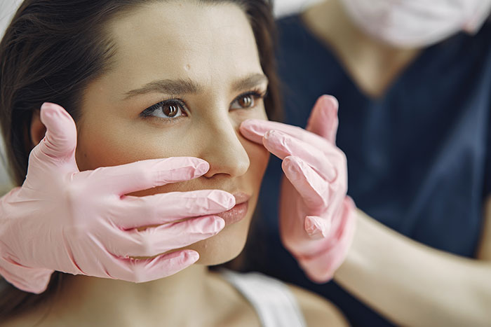Woman receiving facial examination by a professional wearing pink gloves, illustrating botched plastic surgeries people sadly experienced.