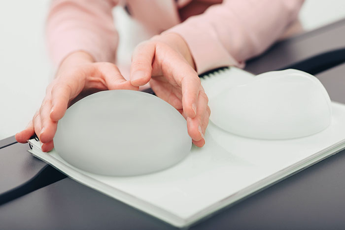Hands holding silicone breast implants on a table, illustrating botched plastic surgery risks and complications.
