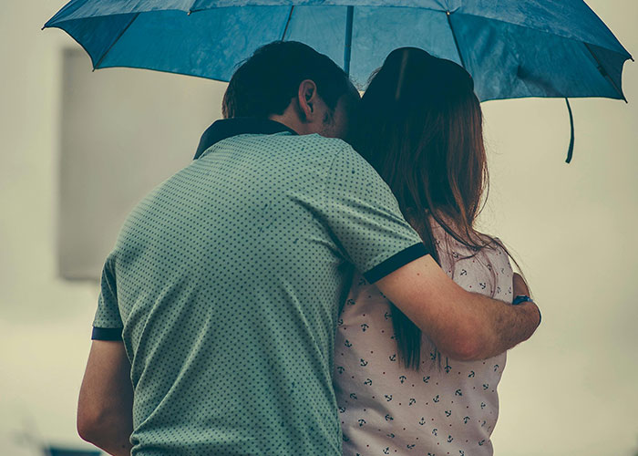 Couple standing closely under a blue umbrella, with the man embracing the woman from behind on a rainy day.