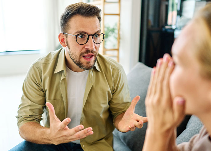 Man with glasses explaining passionately to woman on couch during a heated discussion about chores and rent money.