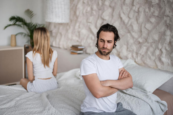 Young man sitting with arms crossed looking upset while his girlfriend turns away on the bed, illustrating boyfriend dating relationships.
