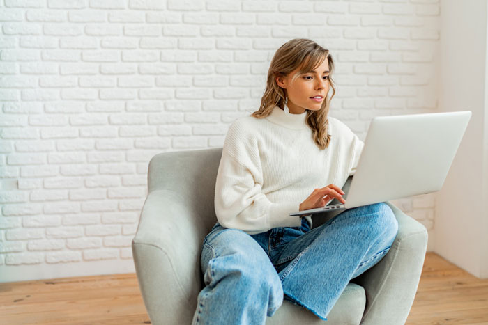 Young woman using laptop sitting in armchair, focused on boyfriend dating relationships through online browsing.