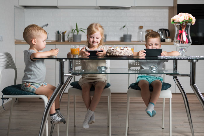 Three children sitting at a kitchen table, focused on tablets, with a cake and drinks nearby, reflecting family expectations.